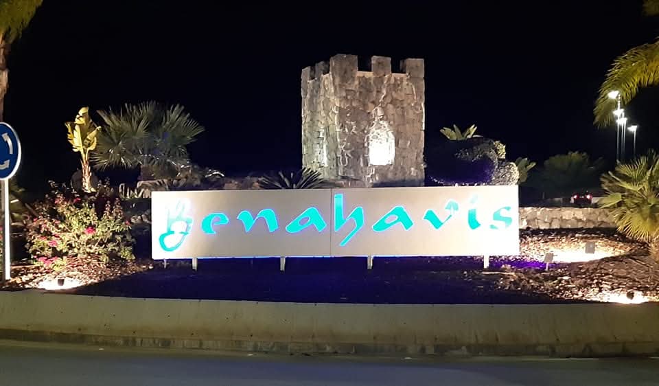 Rotonda iluminada en la entrada de Benahavís con un letrero y una torre de piedra de fondo